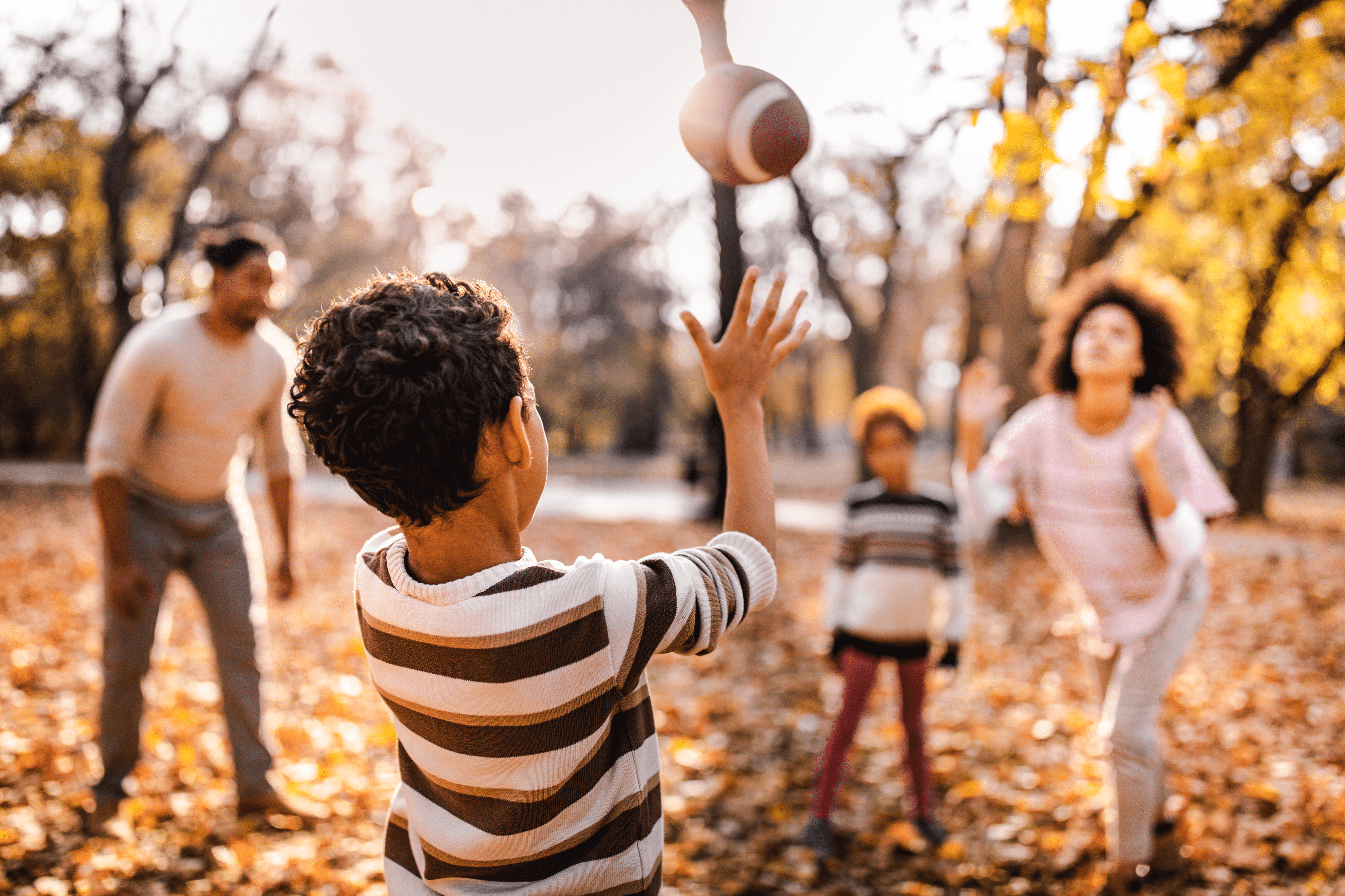 A group of four people (a man, a woman, and two children) toss around a football in an outdoor autumn landscape.