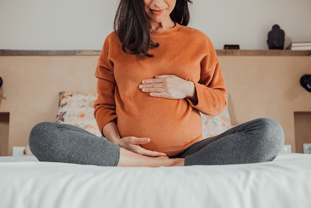 A pregnant person in sweatpants and a sweatshirt sits cross-legged on a bed.
